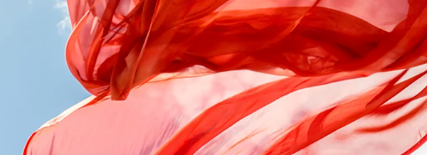 Close-up of sheer red fabric flowing in the wind against a blue sky with scattered white clouds.