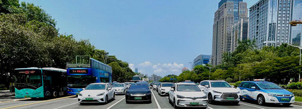 Multiple cars, a police car, and two buses wait at a city junction lined with trees and tall buildings under a clear blue sky.