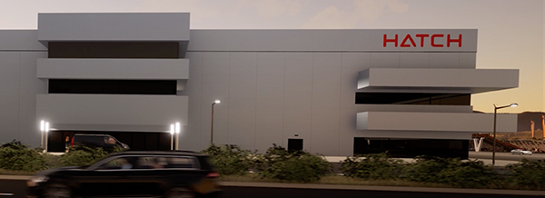 Modern grey building with HATCH in red letters; cars and bushes in front; evening lighting.