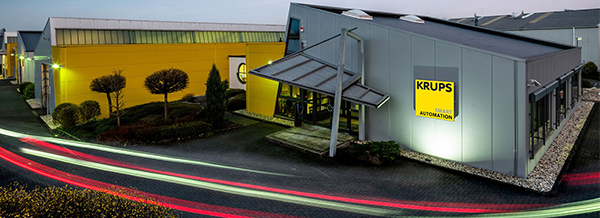 Modern industrial buildings with yellow walls and a sign reading KRUPS, light trails from passing vehicles visible in the foreground.