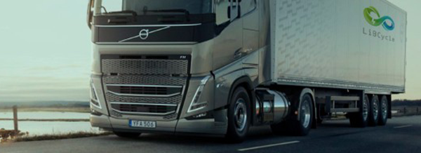 A silver Volvo lorry with a trailer drives on a road by a body of water at sunset.