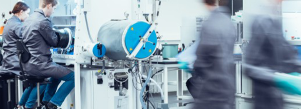 Scientists in lab coats work with technical equipment in a bright, modern laboratory; others walk by in the foreground.